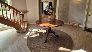 A spacious foyer with a marble floor, a round table with a woven rug, a staircase, and a chandelier.