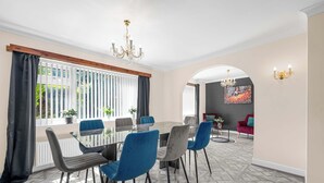 A dining area with a large table, blue and grey chairs, a chandelier, and a view through a window with blinds.