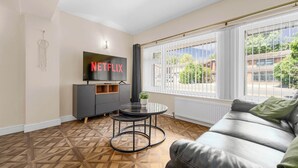 A modern living room with a leather sofa, a round coffee table, a TV mounted on the wall, and a large window with blinds.