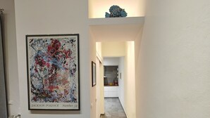 Un couloir moderne avec un tableau abstrait encadré, un canapé noir avec des coussins et un sac de couleur claire posé sur le sol.