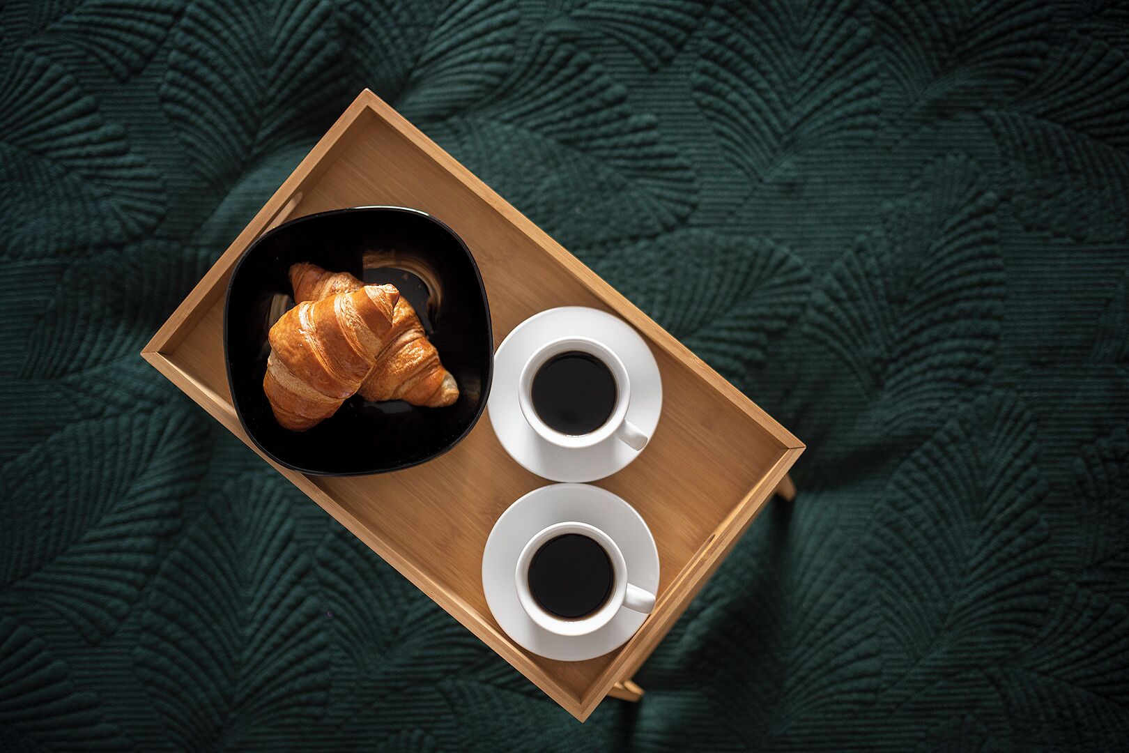 Een houten dienblad met een croissant en twee kopjes koffie op een groen geweven doek.