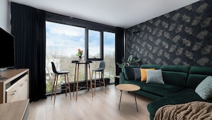 A modern living room with a green sofa, a small round table, and two chairs. There is a large window with a view of trees and a cloudy sky. - EASY RENT Apartments - Brilliant (Lublin)