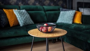 A green sofa with patterned cushions, a round wooden table with a black wire fruit basket, and a patterned wall in the background. - EASY RENT Apartments - Brilliant (Lublin)
