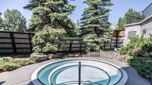 A circular hot tub with a tiled edge, surrounded by a stone border, set in a landscaped outdoor area with trees and a wooden fence. - Stunning Silver Creek 1Br Mtn view penthouse (Canmore)