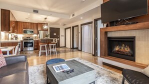 A modern living room with a fireplace, a kitchen in the background, and a dining area with stools. - Stunning Silver Creek 1Br Mtn view penthouse (Canmore)