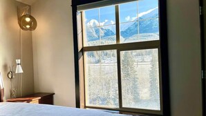 A bedroom with a large window, a bed, a nightstand, and a wall-mounted lamp. - Stunning Silver Creek 1Br Mtn view penthouse (Canmore)