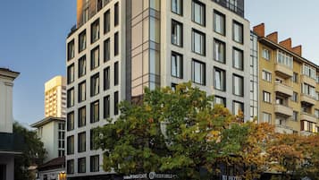 A modern hotel with a prominent 'BI HOTEL' sign, multiple floors, and a street view with trees and storefronts.