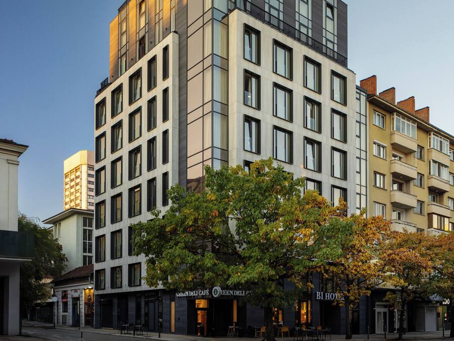 A modern hotel with a prominent 'BI HOTEL' sign, multiple floors, and a street view with trees and storefronts.