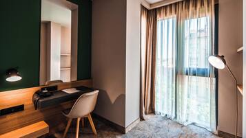 Quarto de hotel com escrivaninha, cadeira, espelho e uma janela grande com cortinas.