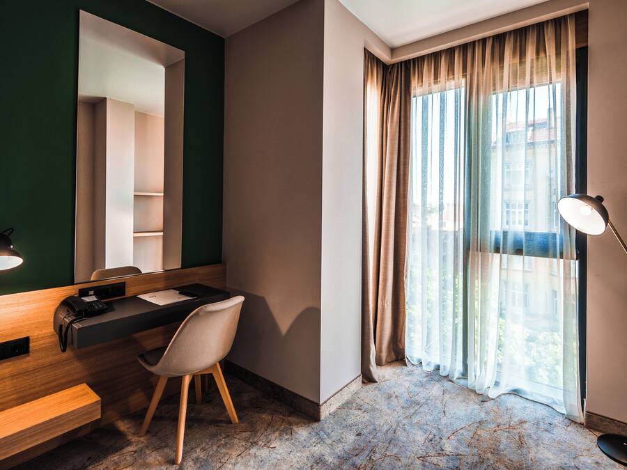 Quarto de hotel com escrivaninha, cadeira, espelho e uma janela grande com cortinas.