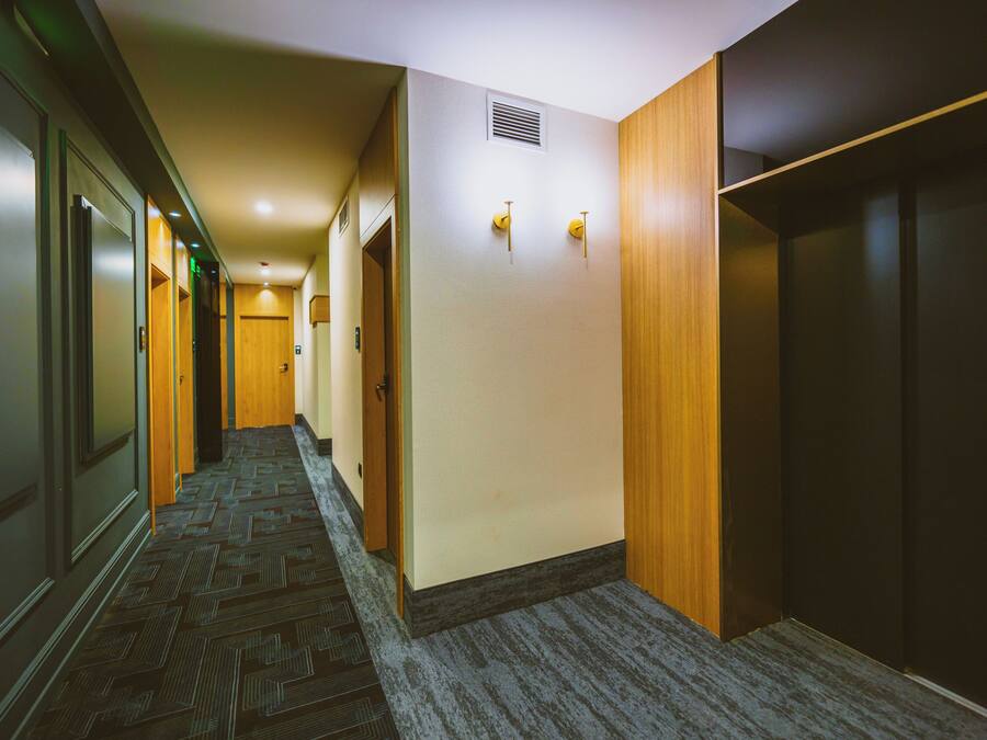 A hotel hallway with carpeted floors, wooden doors, and a ventilation system on the wall.