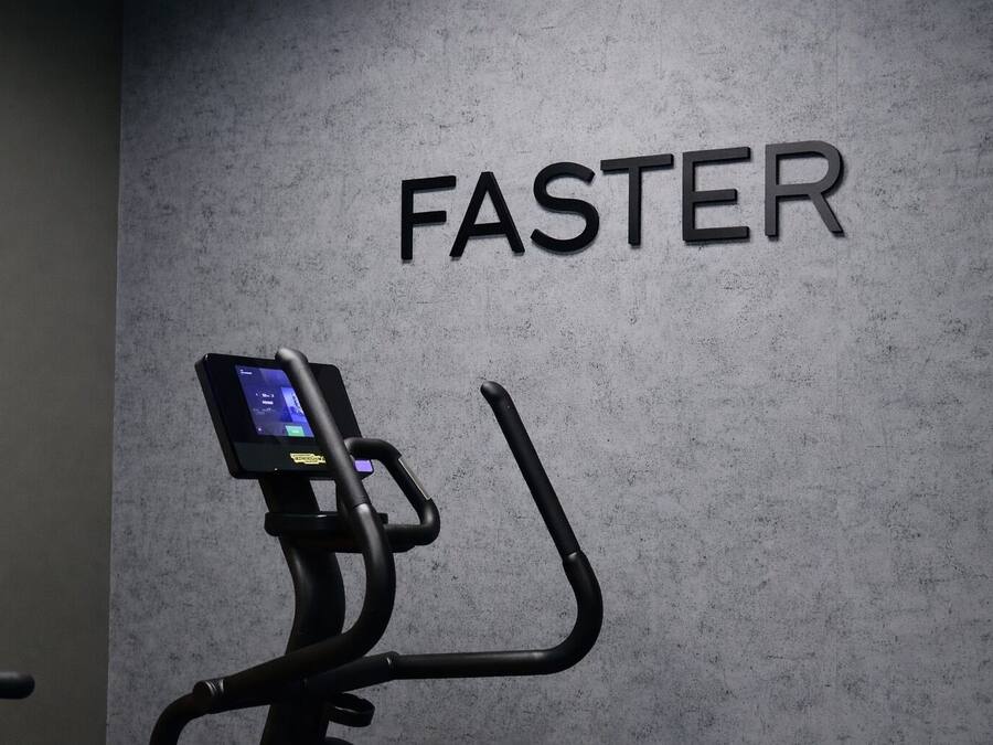 Una bicicleta estática con pantalla digital y manubrios, ubicada frente a una pared donde se lee la palabra 'FASTER'.