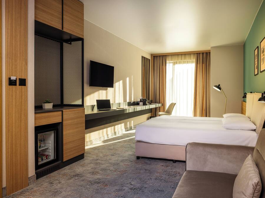 Habitación de hotel con cama, sofá, escritorio con computadora, televisor y ventana con cortinas.