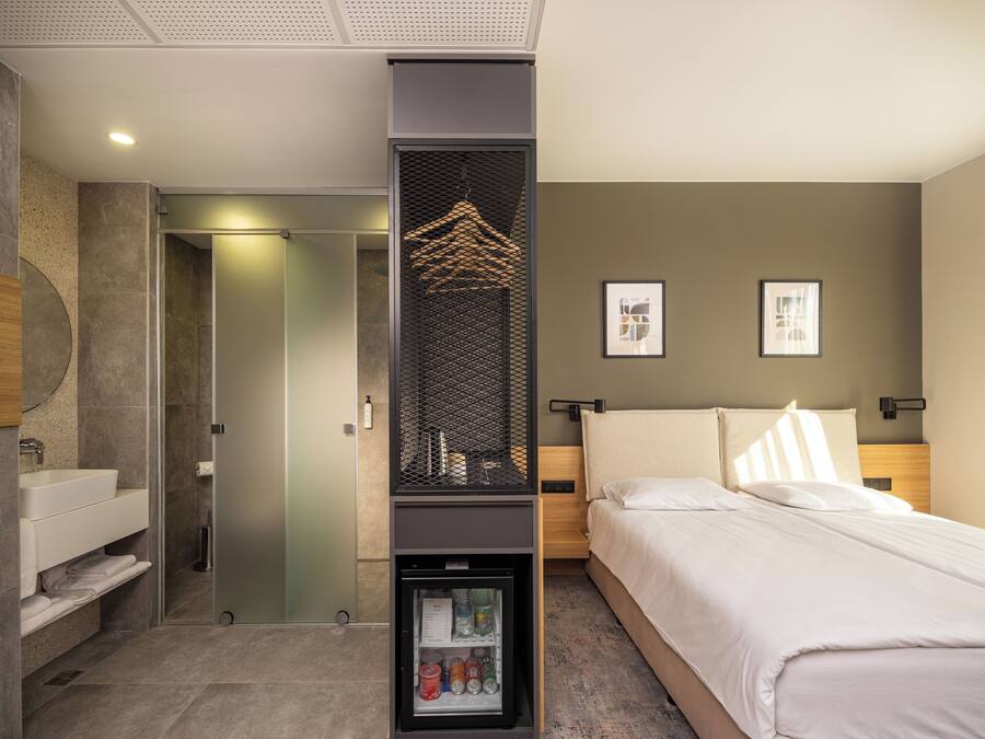 Una habitación de hotel moderna con una cama, un minibar integrado, zona de ducha y baño con lavamanos y espejo.