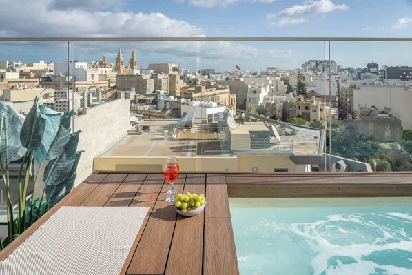 A rooftop terrace with a pool, a table with a bowl of fruit and a glass of red drink, and a city view.