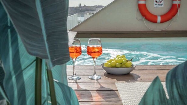 A poolside view with two glasses of orange beverage, a bowl of grapes, and a life ring on a wooden deck. - Penthouse, pool & Valletta views | ROP by Homega (Sliema)