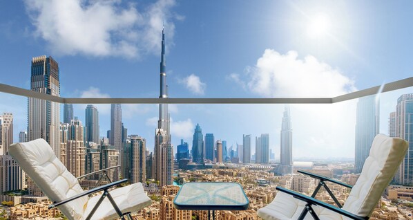 Un balcón con dos sillas blancas acolchadas y una mesita, con vistas a un paisaje urbano de edificios altos y cielo despejado.