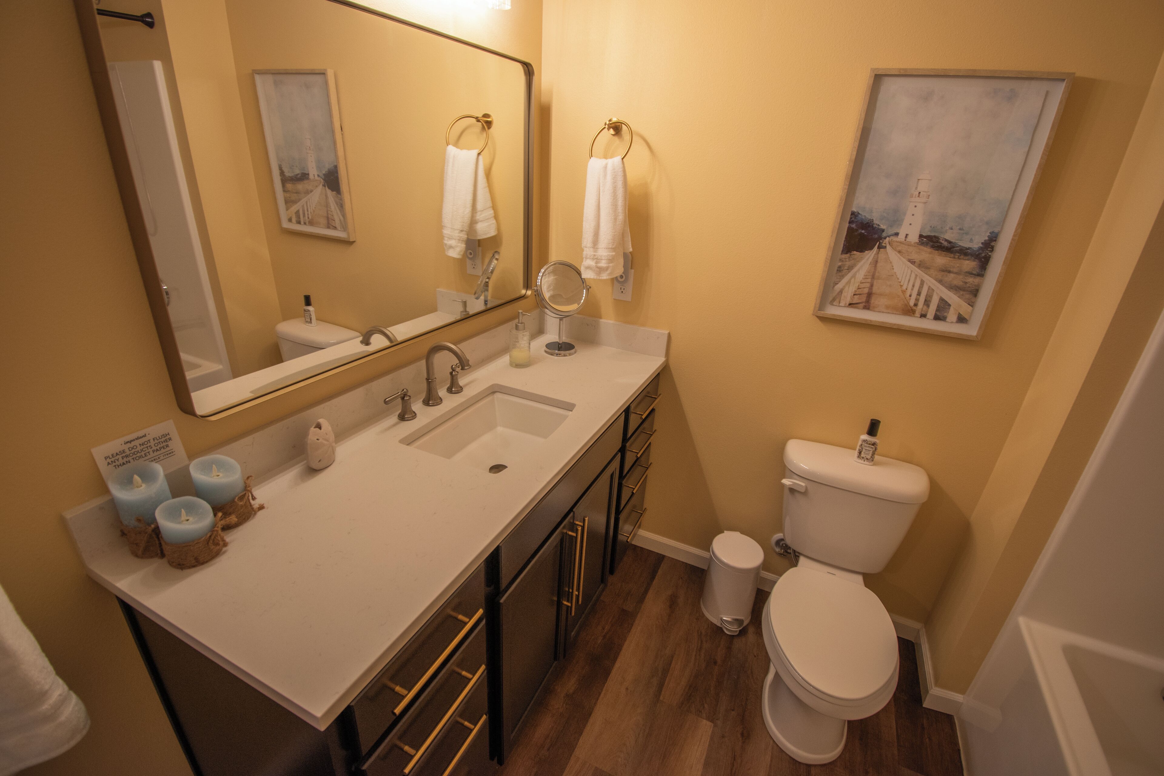 A bathroom with a white sink, mirror, and toilet, wooden flooring, and a painting on the wall.