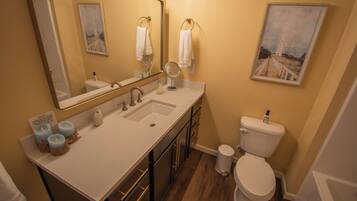 A bathroom with a white sink, mirror, and toilet, wooden flooring, and a painting on the wall.