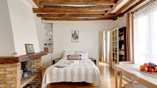 A cozy bedroom with a brick fireplace, a bed with striped bedding, a wooden ceiling, and a bookshelf. - Charming 1BR near Eiffel Tower & Les Invalides (Paris)