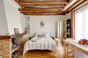 A cozy bedroom with a brick fireplace, a bed with striped bedding, a wooden ceiling, and a bookshelf.