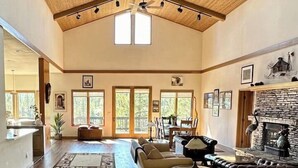 A spacious living room with a high ceiling, wooden beams, a fireplace, and a large window. - The Alpine Vista | Pets | Couples | Golf | Nakoma (Portola)