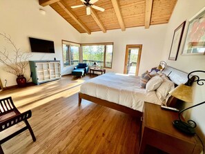 A bedroom with a large bed, wooden flooring, a ceiling fan, a TV, and a window with a view of trees. - The Alpine Vista | Pets | Couples | Golf | Nakoma (Portola)