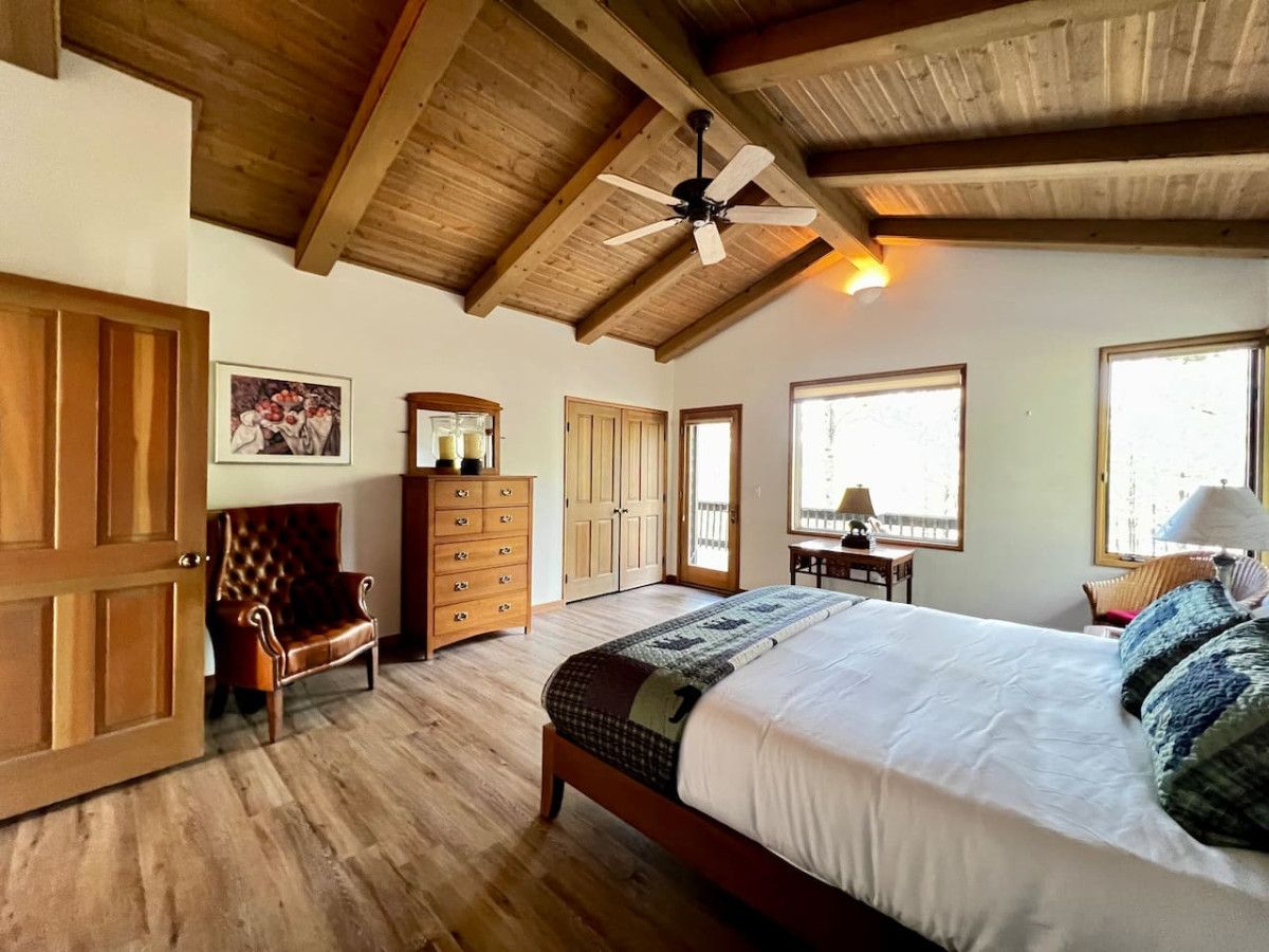 A bedroom with a large bed, wooden ceiling beams, a ceiling fan, and a view of the outdoors through a window.