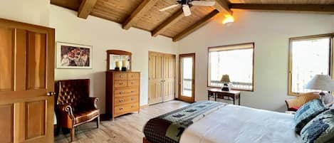 A bedroom with a large bed, wooden ceiling beams, a ceiling fan, and a view of the outdoors through a window.