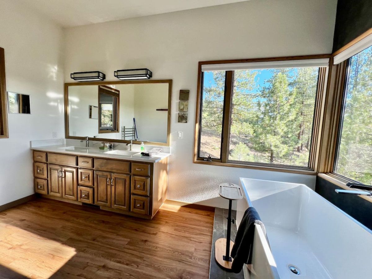 A modern bathroom with a large mirror, wooden cabinets, a bathtub, and a window with a view of trees.