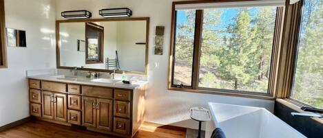 A modern bathroom with a large mirror, wooden cabinets, a bathtub, and a window with a view of trees.