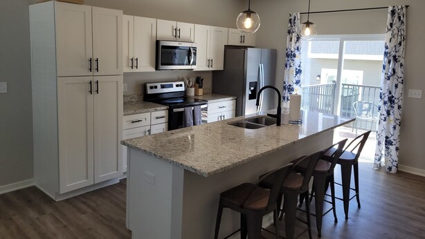 A modern kitchen with white cabinets, a stainless steel refrigerator, a microwave, and a granite countertop. There are three bar stools at the island. - Stylish Family Townhome with Relaxing Deck Views (Omaha)
