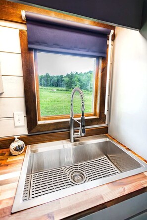 A kitchen with a stainless steel sink, a window with a view of a green landscape, and wooden cabinetry.