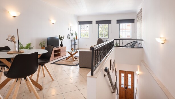 A spacious living area with a dining table, black chairs, a sofa, and a TV. There are windows with blinds, a staircase, and a railing. - Oasis apartment - City center by HD (Faro)