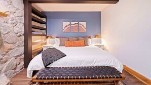 Un dormitorio con pared de piedra, una cama con ropa de cama blanca, dos lámparas de pared y un banco con asiento de mimbre.
