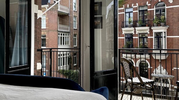 A bedroom with a large bed, a view of a brick building, a balcony with a chair and table, and a hanging light fixture.