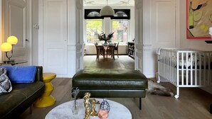 A living room with a green ottoman, a yellow side table, a white crib, and a dining area with a table and chairs.