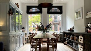 A dining area with a wooden table and chairs, a vase with flowers, and a view of buildings through large windows.