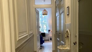 A hallway with white walls and a marble floor leading to a room with a window, a hanging light fixture, and a sink.