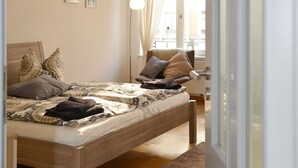 Une chambre à coucher avec un lit en bois, une grande fenêtre avec des rideaux, un luminaire suspendu et une petite table de chevet.