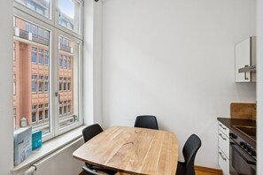 Moderni keittiö, jossa on puinen pöytä ja tuolit, ikkuna, josta avautuu näkymä punaiseen rakennukseen, sekä valkoinen kaappi.