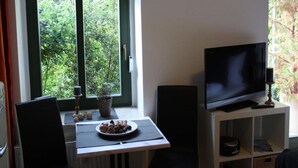 A room with a TV on a stand, a dining table with a plate of food, and a window with a view of greenery. - Apartment-Ensuite with Shower-Terrace near Bayerischer (Leipzig)