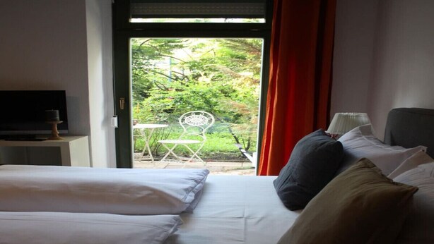 A hotel room with a bed, pillows, a TV, and a view of greenery outside. - Apartment-Ensuite with Shower-Terrace near Bayerischer (Leipzig)
