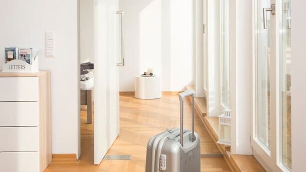 Une chambre d’hôtel moderne et lumineuse, avec un parquet, une grande fenêtre et une valise posée au centre.