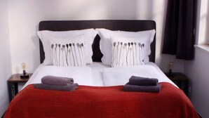 Ein ordentlich bezogenes Bett mit weißer und grauer Bettwäsche, einer roten Decke und zwei gefalteten Handtüchern auf der Seite.