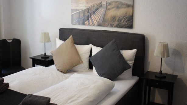 A hotel room with a large bed, two bedside tables with lamps, and a painting of a sunset over a pier. - Apartment-Ensuite with Bath-City view (Leipzig)