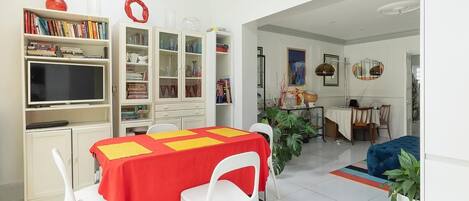 A modern living room with a dining area, featuring a red table, white chairs, a bookshelf, and a television.