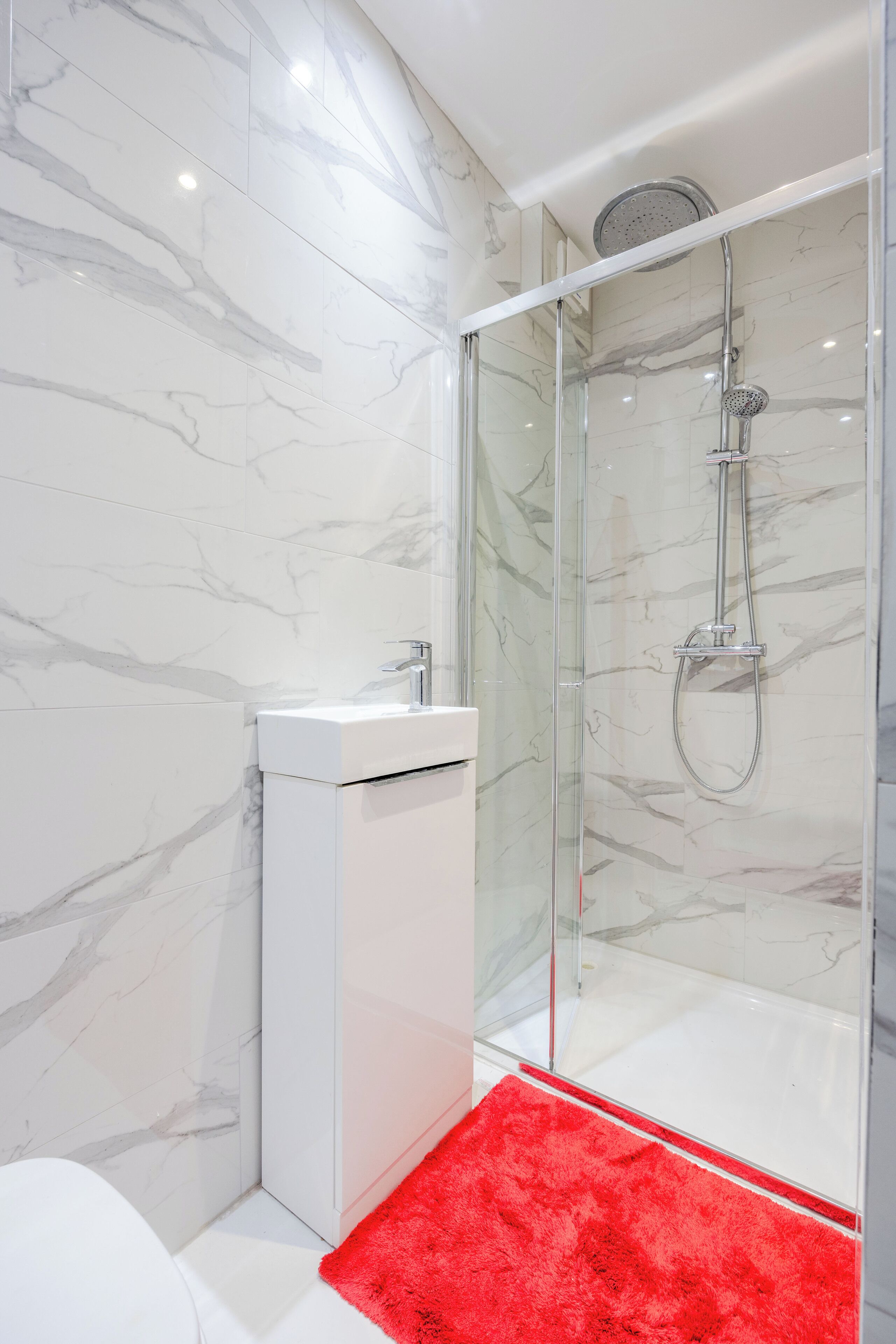 A modern bathroom with a glass-enclosed shower, a white sink, and a red bath mat.