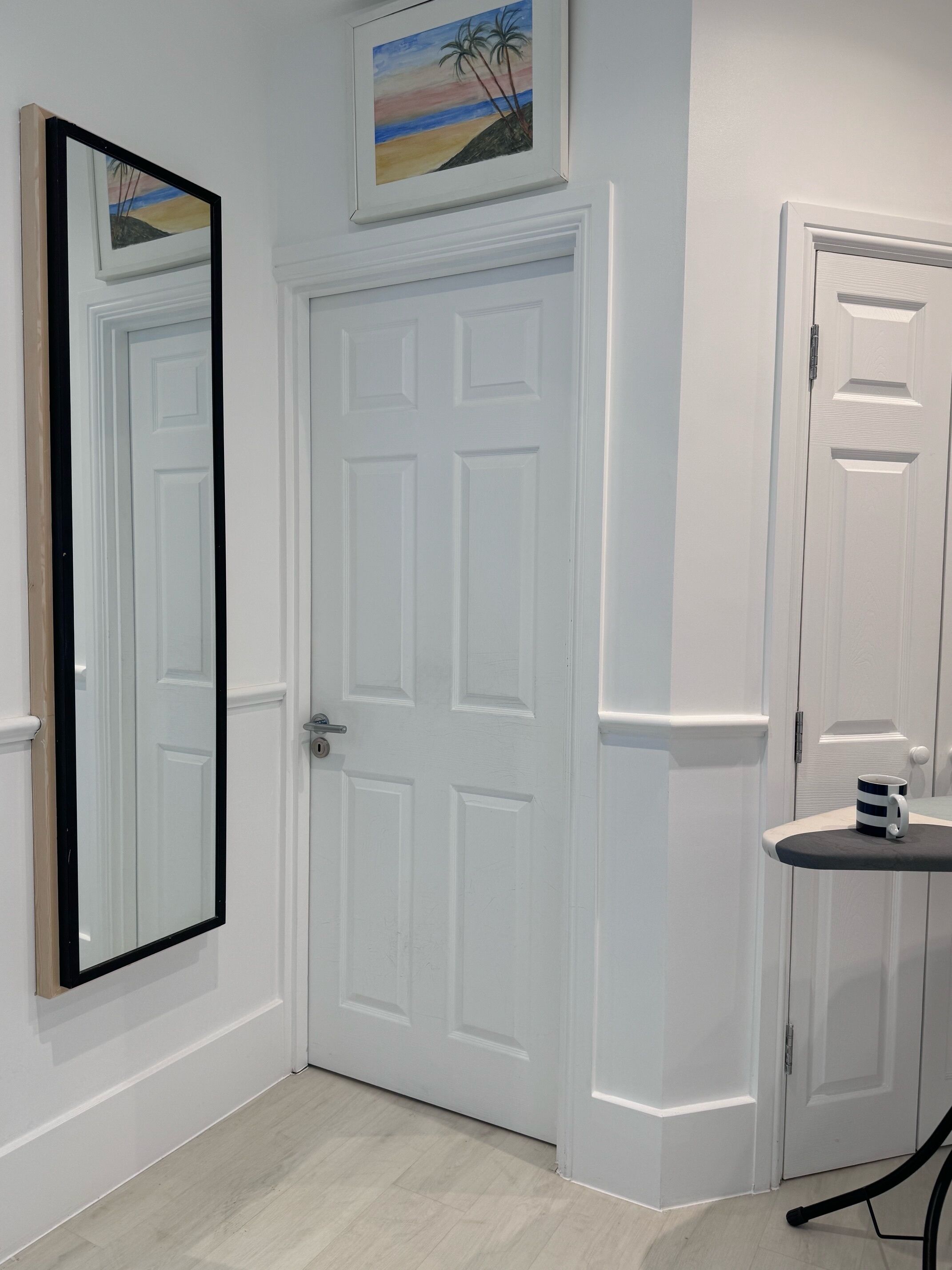 A white door with a keyhole, a framed painting above it, and a small table with a mug.