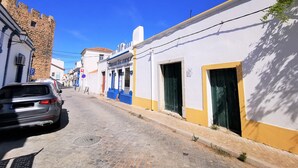 A cobblestone street with buildings on either side, including a white building with a blue door and a stone building with a red door.